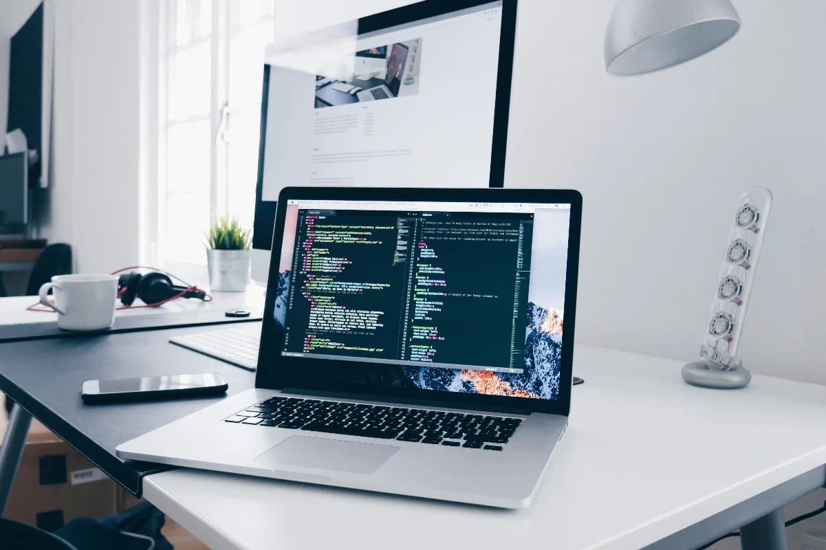 The image depicts a laptop on a white desk, displaying lines of code on its screen. The laptop is open and positioned at an angle, with the keyboard facing the bottom-right corner of the image; it sits on a white desk alongside various office supplies and a monitor.