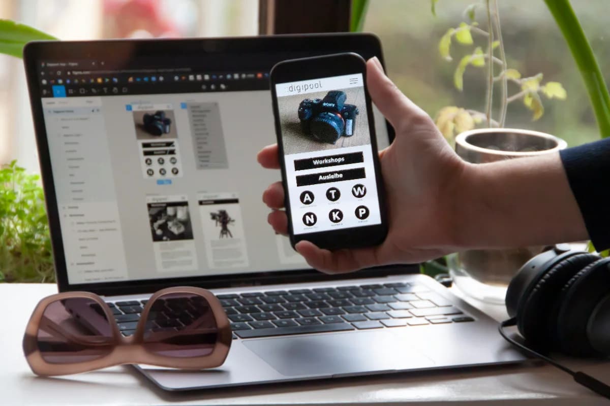 The image depicts a person's hand holding a smartphone with a website on the screen, positioned in front of an open laptop displaying a similar website. The scene is set on a white desk with various items, including sunglasses and headphones.